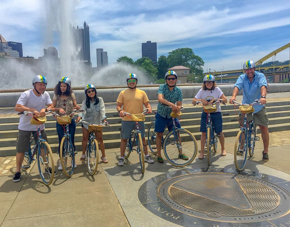 Photo shows a group of visitors on bicycles at Point State Park, with the fountain and city skyline in the background