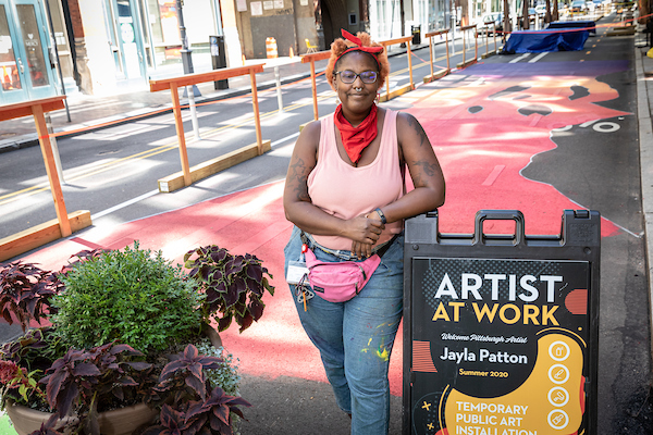 Photo shows artist Jayla Patton standing in front of a red, orange, and purple painted Penn Avenue in Downtown Pittsburgh