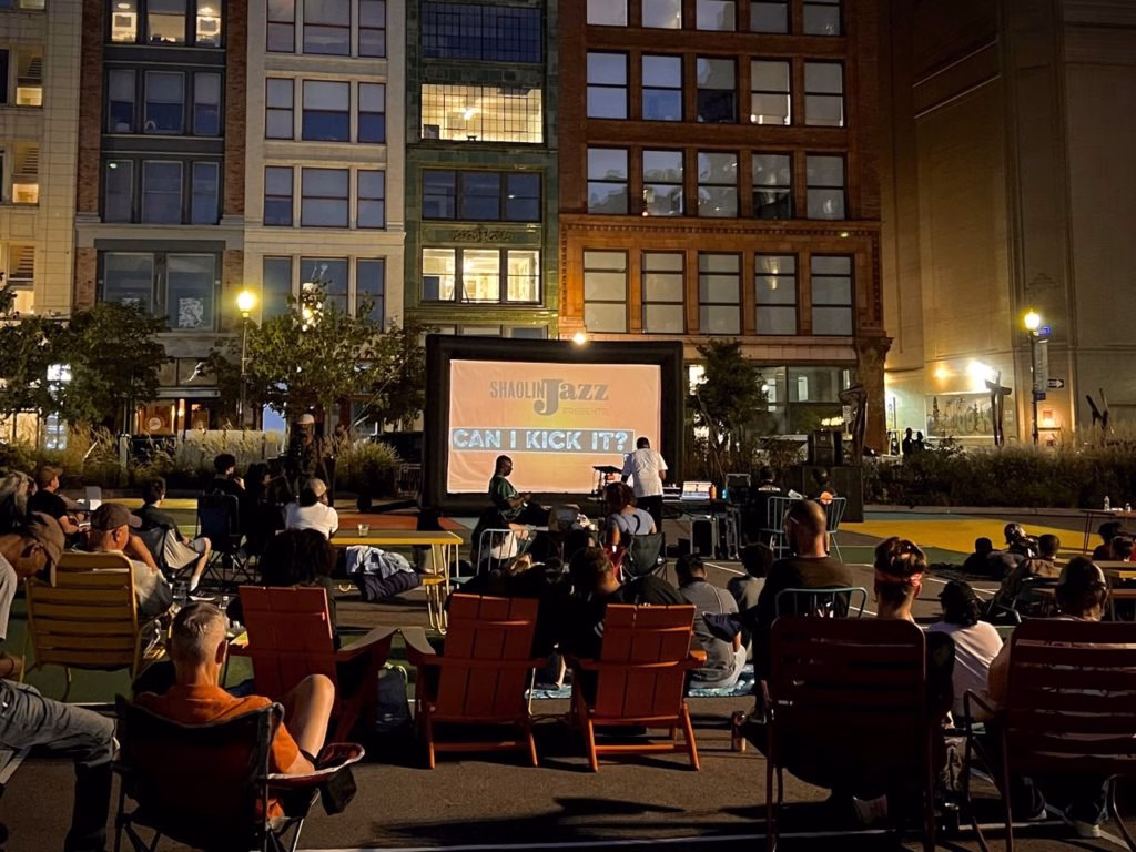 A crowd of people sitting in chairs look up to a screen with the the event title "Shaolin Jazz" projected onto it.