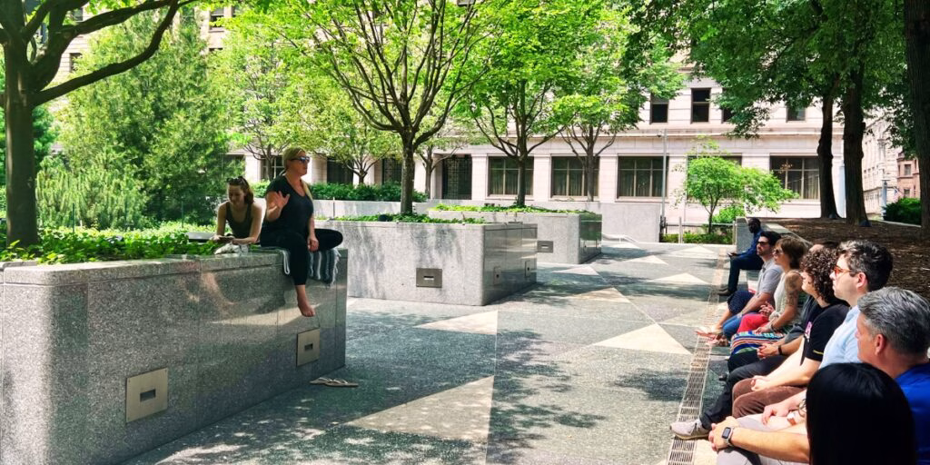 A woman speaking on a ledge in a park to a group of people on a bench.