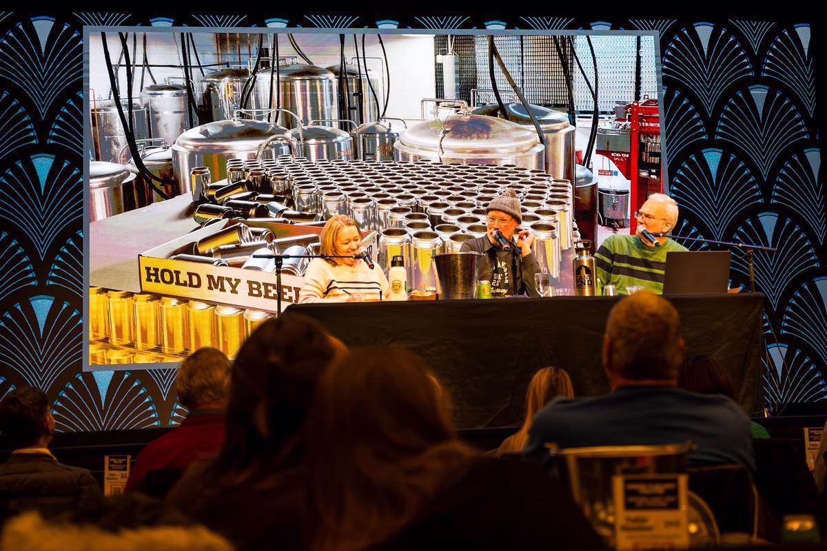 A three mic'd up people on a stage with a massive canning back drop for a brewery. A thirsty crowd looks on.