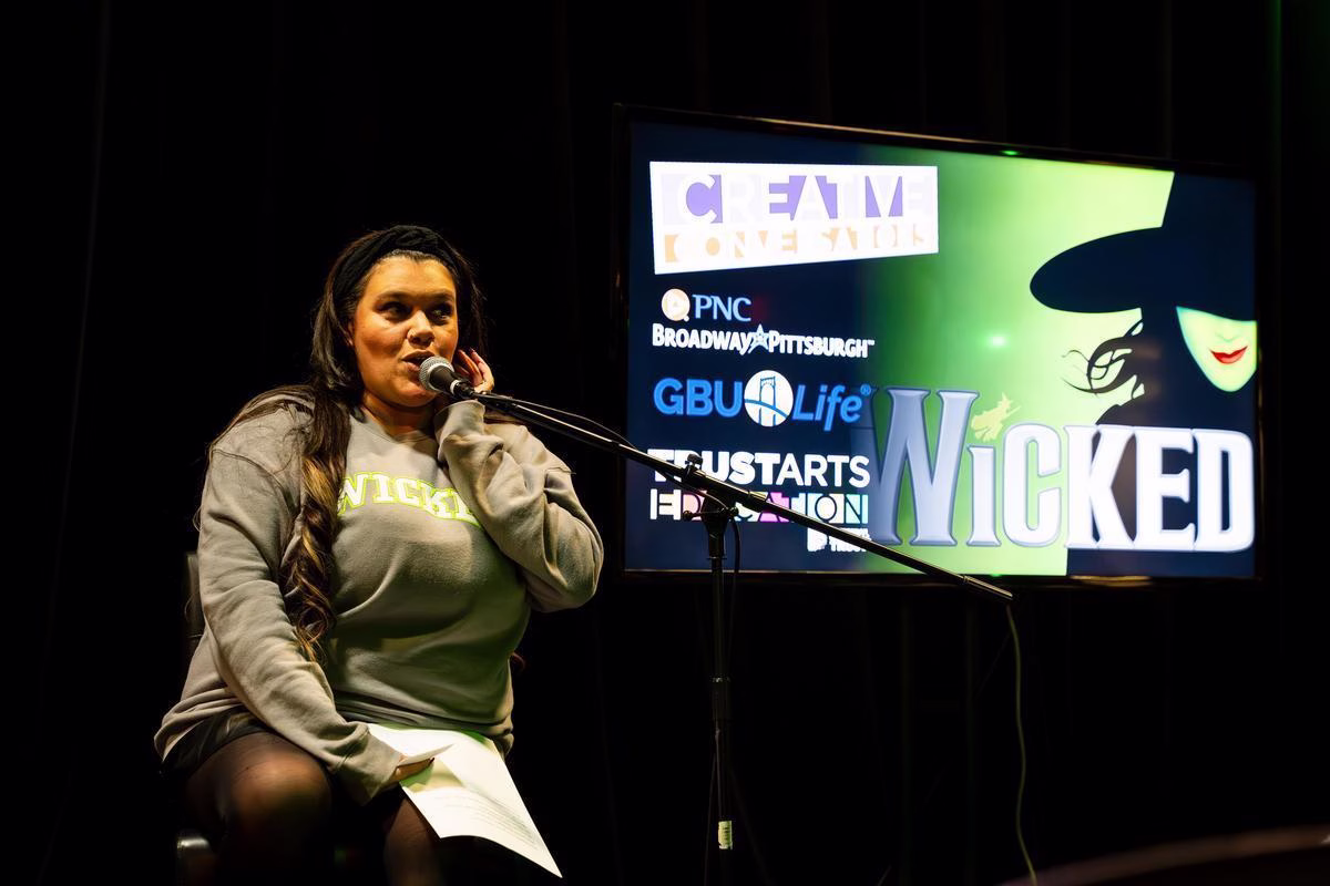A woman speaking into a mic on stage wearing a wicked sweater, sitting in front of a TV with a Wicked poster displayed.