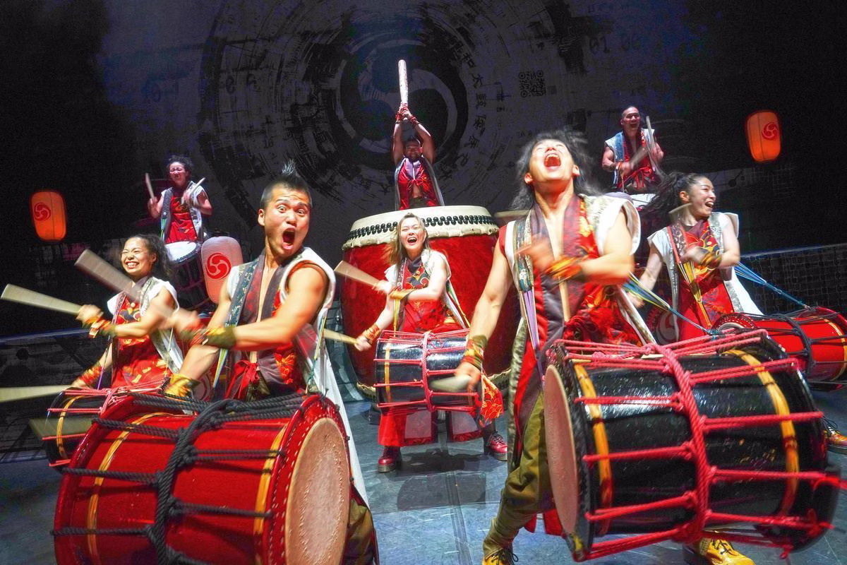 Eight performers on stage banging on drums with excited looks on their faces.