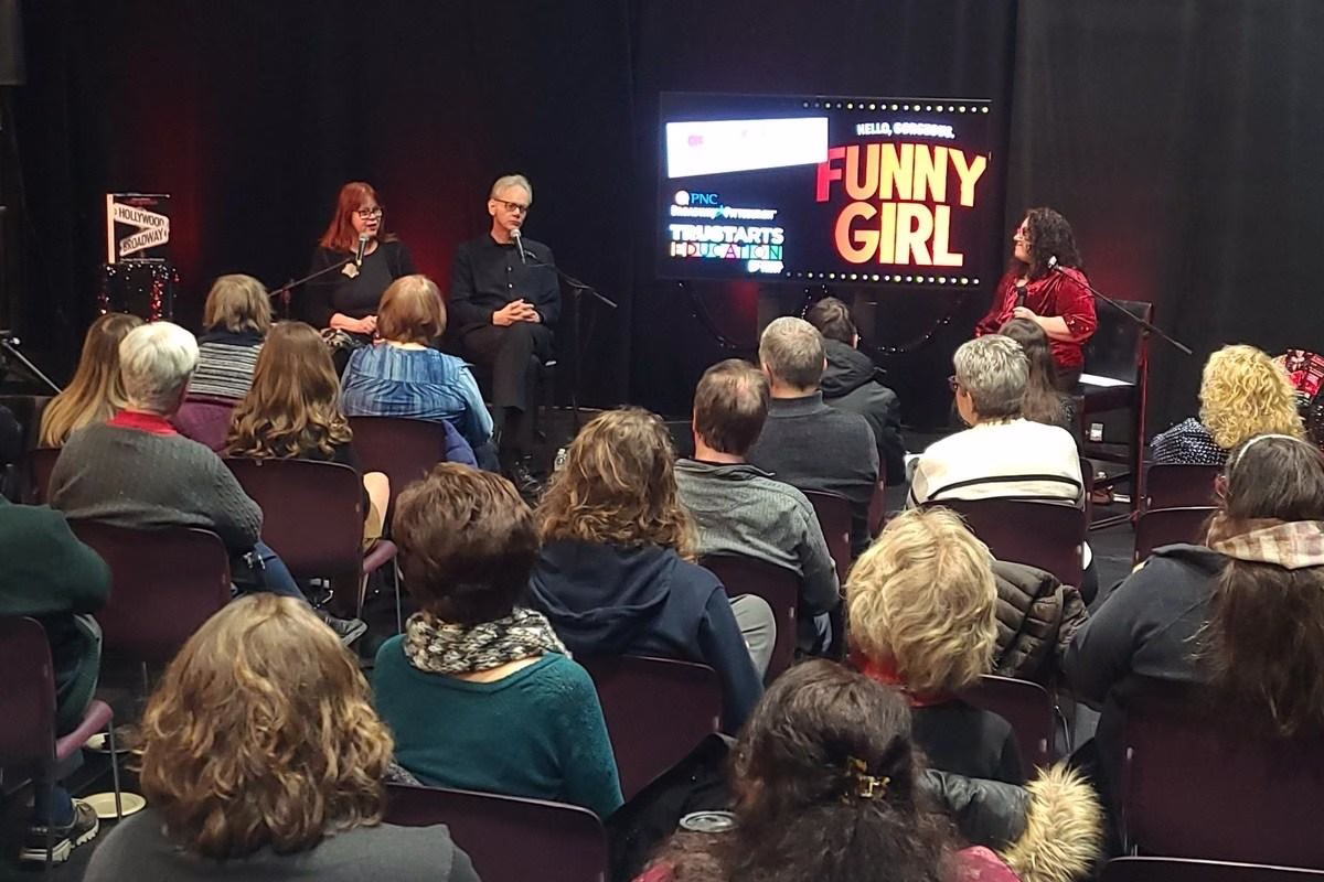 Three people discuss Funny Girl on stage while a an audience of folks eagerly listen.